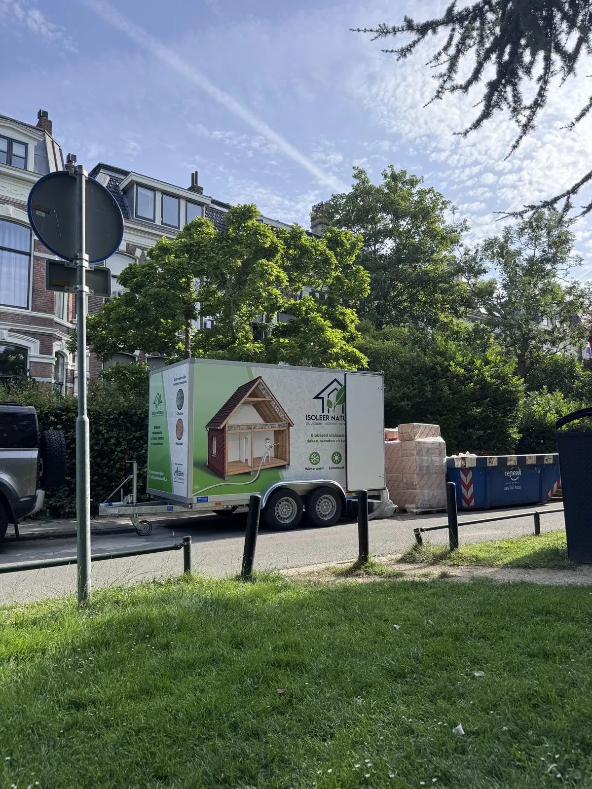 De aanhanger van Isoleer Natuurlijk met inblaasmachine, geparkeerd in een groene straat met herenhuizen